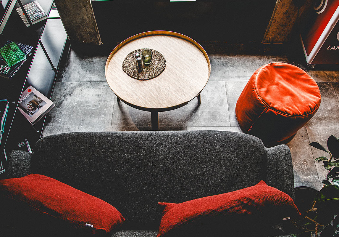 Runder Holztisch mit Salz- und Pfefferstreuer und Glas auf grauem Boden vor dunkelgrauem Sofa mit roten Kissen und rotem Sitzhocker in the store der Serviced Apartments in Herzogenaurach