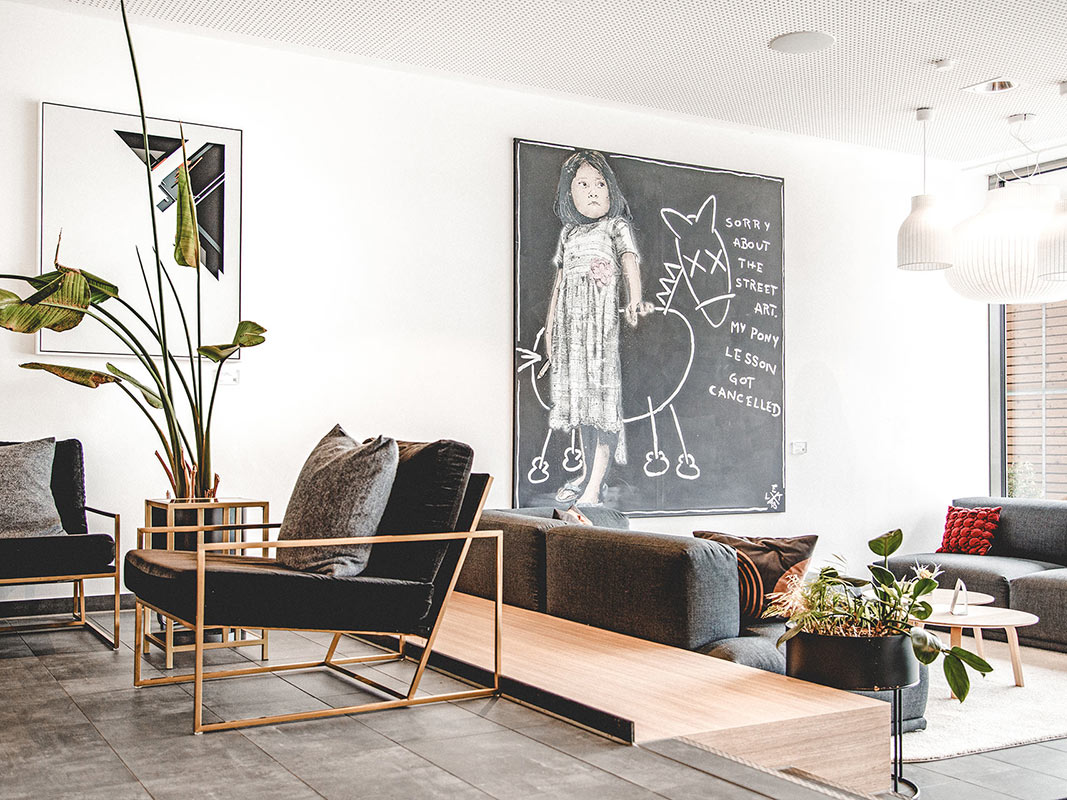 Moderner Aufenthaltsraum mit schwarzen Sesseln, grauem Sofa, großem Pflanzenarrangement und Wandbild mit Kind und gezeichnetem Tier mit Text