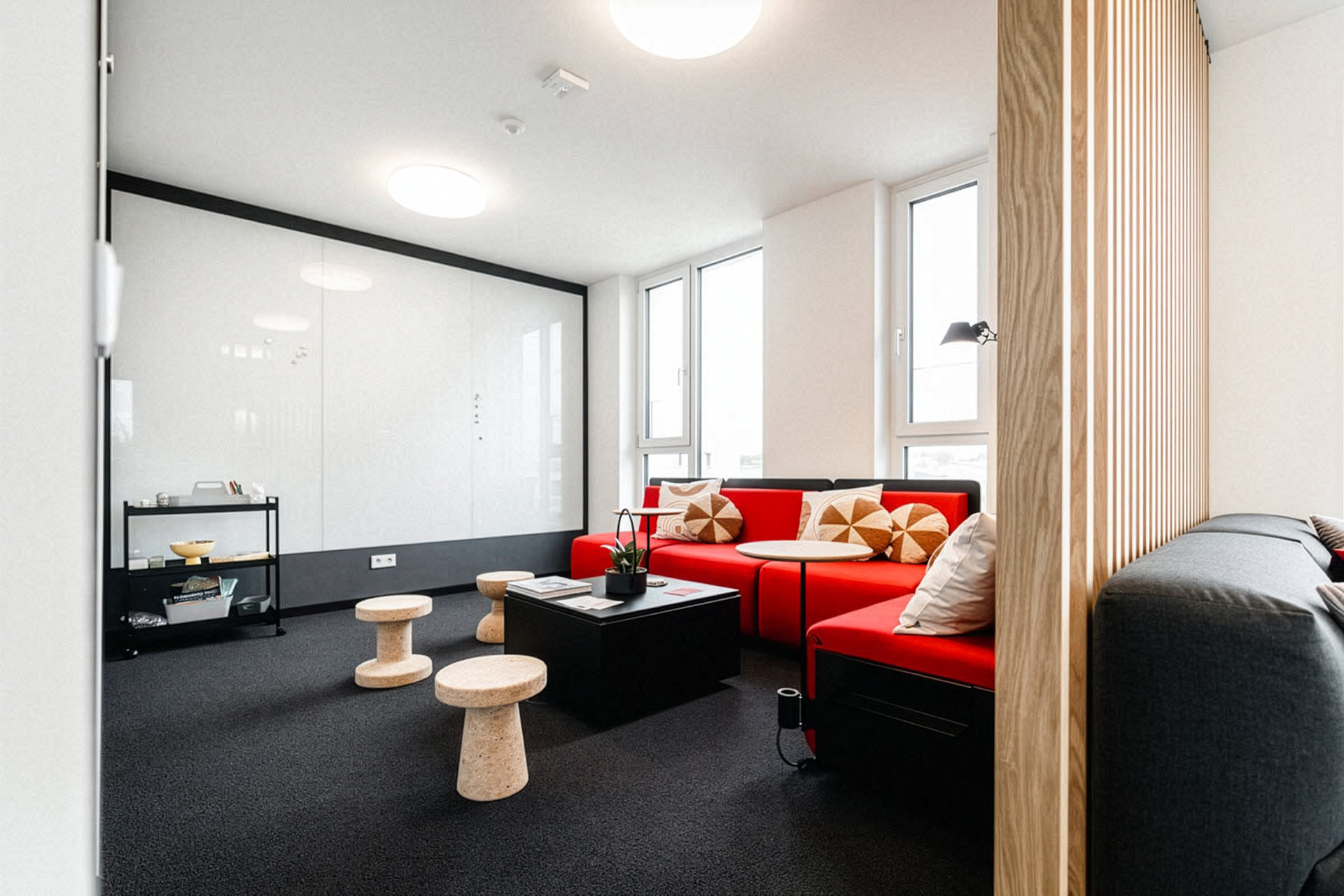 Lounge-Ecke mit rotem Ecksofa und gemusterten Kissen, schwarzem Couchtisch, runden Korkhockern, großer Glaswand als Whiteboard und Holzlamellen-Trennwand