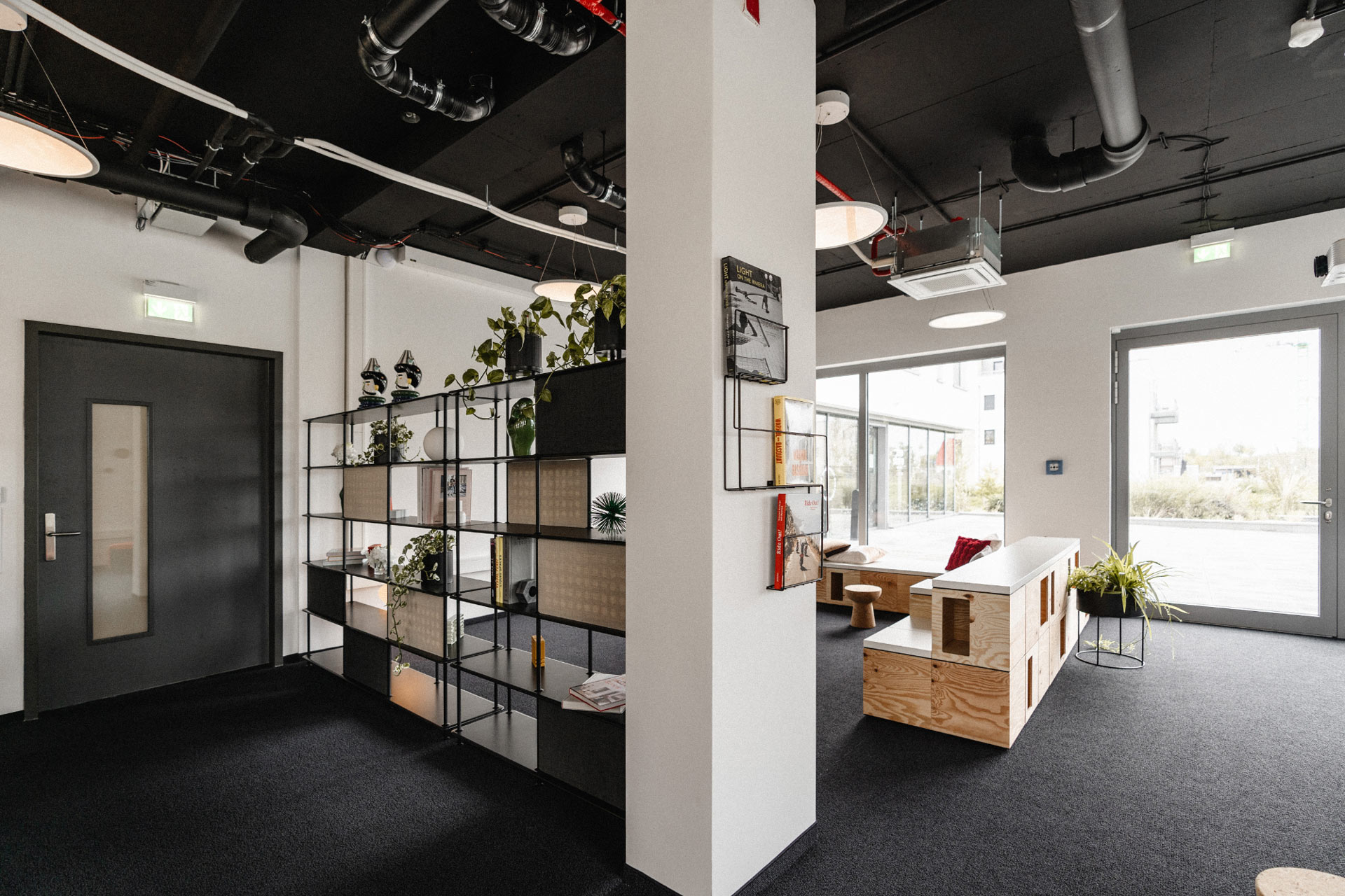 Offener Bereich im Industrial-Design mit schwarzer Sichtdecke und Rohrleitungen, offenem Regalsystem mit Pflanzen und Deko-Objekten als Raumteiler, sowie einer Loungezone mit Holz-Sitzmöbeln und großen Fenstern zum Außenbereich